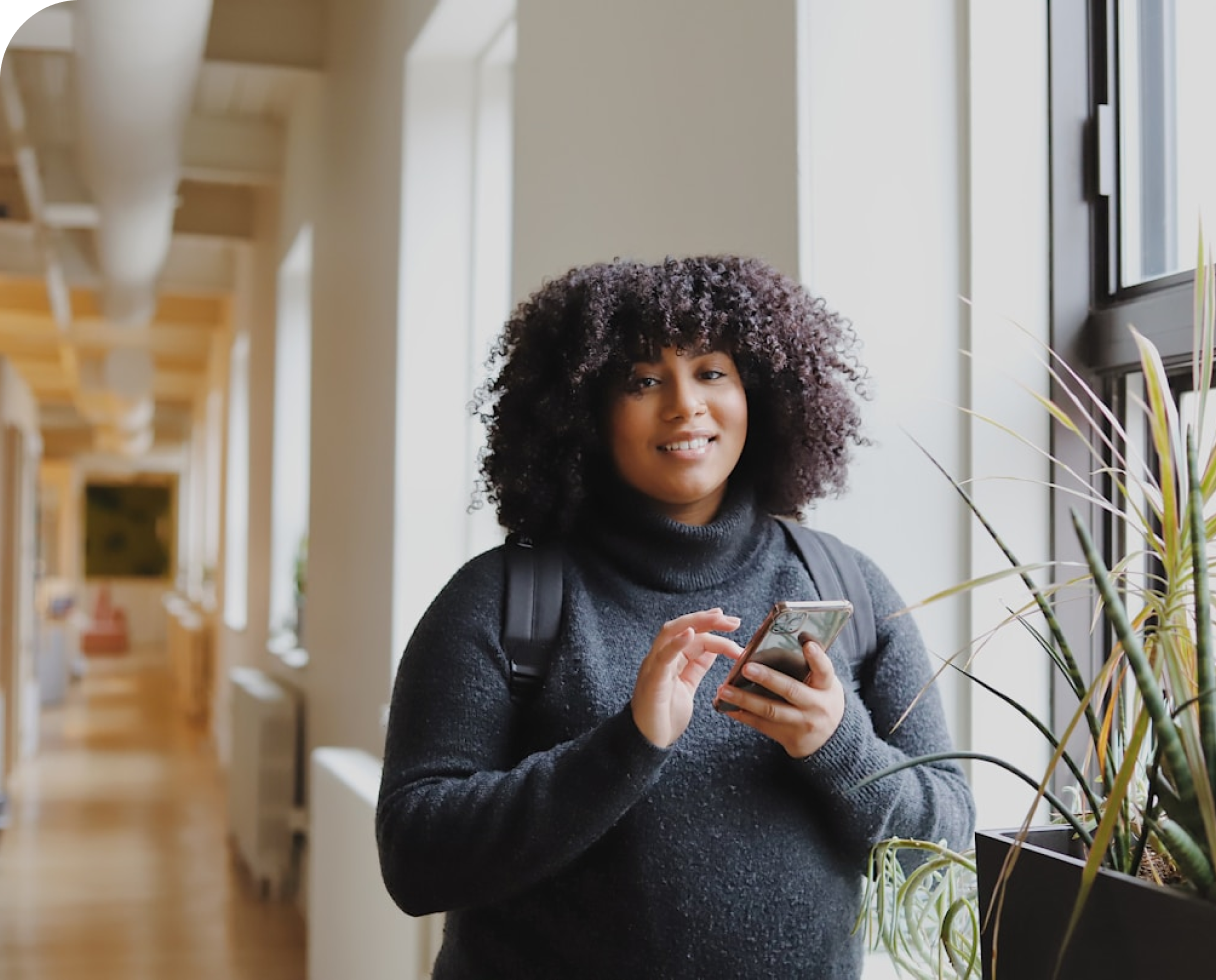 Woman with black curly hair wearing a backpack in a hallway holding and about to tap a phone
